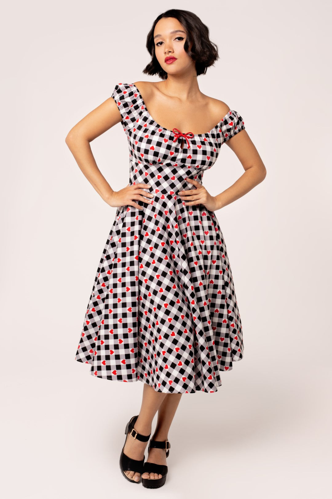 A model wearing a 50's style black and white gingham dress with red hearts, off-the-shoulder neckline, and a red bow on the keyhole front.